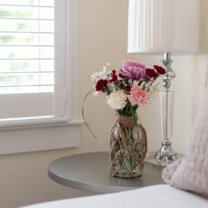 Partial view of a white bedroom with taupe accents. Silver bed side table with lamp and vase of flowers