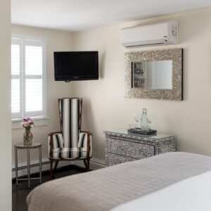 White bedroom with taupe accents. Black and white chair, bureau, small table, tv on the wall as well as a mirror and several pictures.