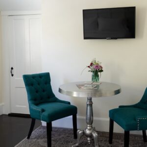 A silver table with a vase of flowers, 2 teal colored chairs, a tv on the wall and a full length mirror