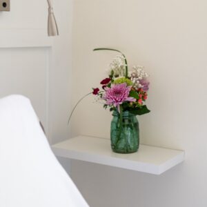 A corner of a white be with a small shelf beside it holding a vase of flowers