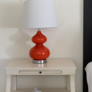 An off white night stand with an orange lamp an a sea shell on one shelf