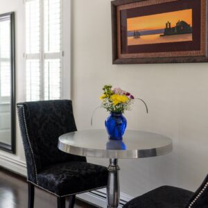 Silver table with a blue vase full of flowers, 2 black chairs, a painting with orange tones on the wall and a tall mirror framed in black