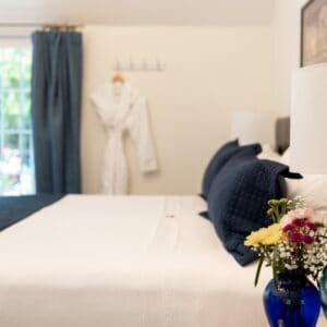 Bedroom with white bed and blue accents. In the forground is a vase of flowers and turquoise table lamp.