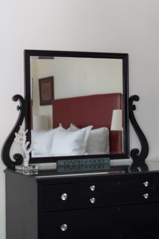 The reflection in a bureau mirror of a bed with a red leather headboard and white pillows is