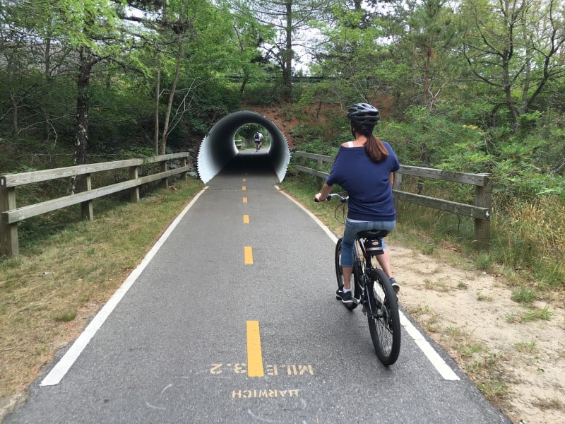 A woman in a blue shirt and jeans wearing a helmet riding a bicycle on the Cape Cod Rail Trail