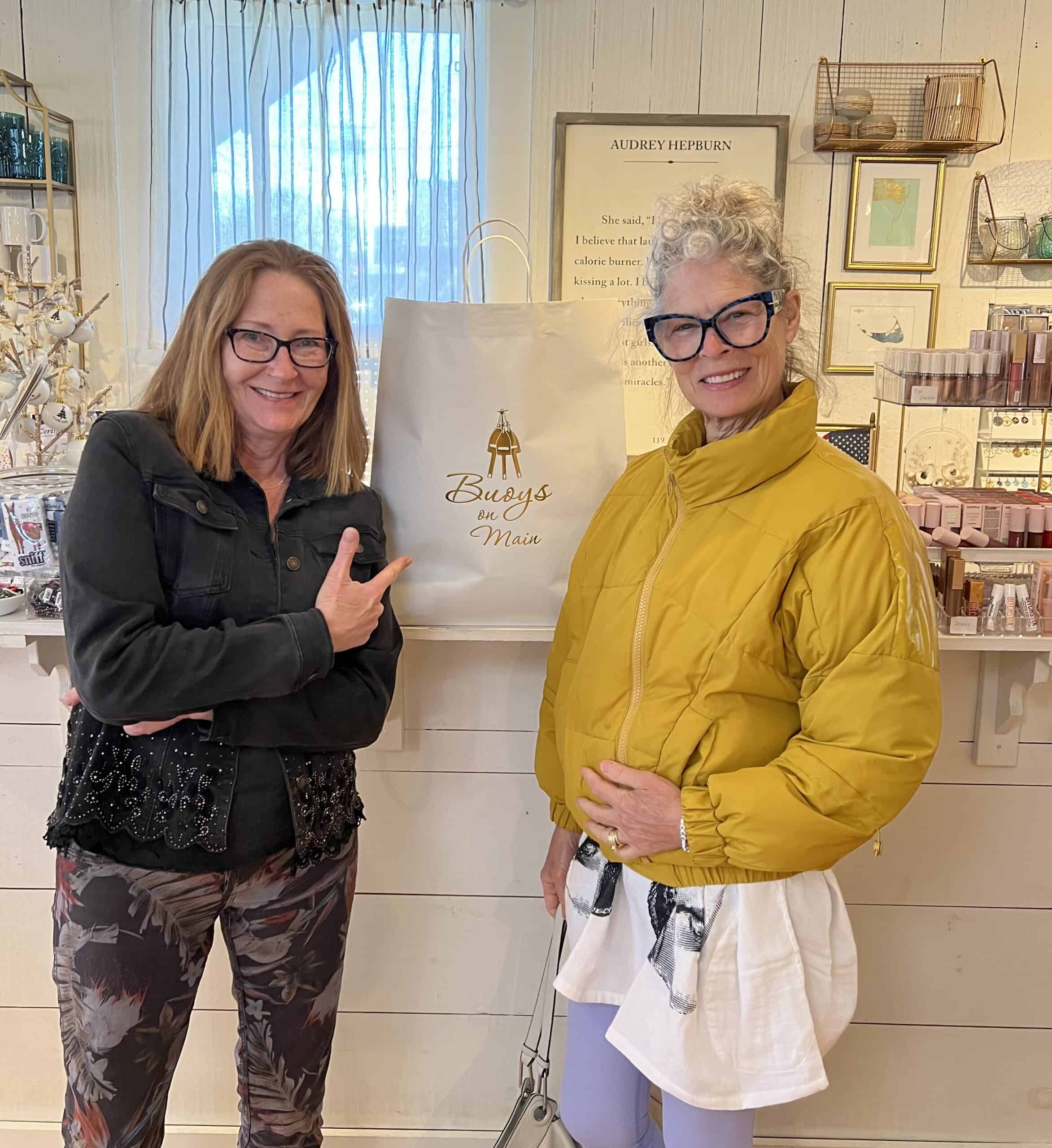 Two women in a store.  One is blond with red eye glasses and wearing a yellow puffy coat.  The other is wearing black eye glasses, a black bejeweled jacket and is pointing at a white shopping bag.